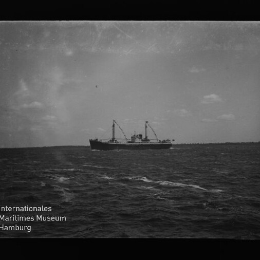 Frachtschiff mit zwei Ladebäumen auf offener See, weiter Horizont und Museumswasserzeichen unten links