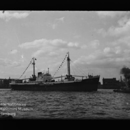 Frachtschiff mit Flaggen im Hafen, dahinter Industriegebäude und rechts ein rauchender Schlepper; Museumssignet links.