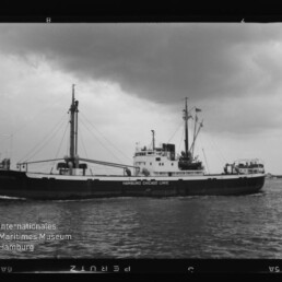 Schwarzweißfoto eines Frachtschiffs der Hamburg-Chicago-Linie mit Masten und Aufbauten unter bewölktem Himmel.