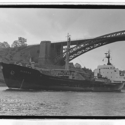 Schwarzweißfoto eines Frachtschiffs unter einer Bogenbrücke, Bäume am Ufer und Museumswasserzeichen unten links.