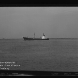 Schwarzweißfoto eines Frachtschiffs auf ruhiger See vor flachem Horizont, mit dem Schriftzug Internationales Maritimes Museum Hamburg.