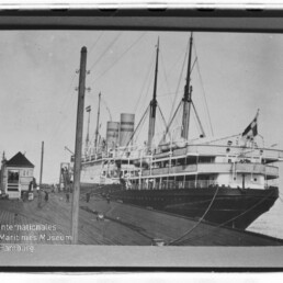 Großes Dampfschiff mit zwei Schornsteinen und Masten am Kai, Personen auf Pier; Wasserzeichen Int. Maritimes Museum Hamburg