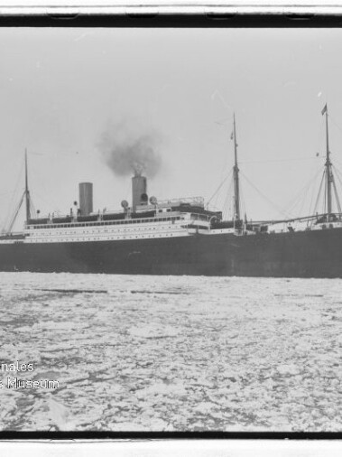 Historisches Dampfschiff mit schwarzem Rumpf und rauchender Schornstein fährt durch eisbedecktes Meer; Museumslogo unten links.