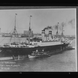 Großes historisches Passagierschiff mit zwei rauchenden Schornsteinen, begleitet von Schleppern im Hafen.