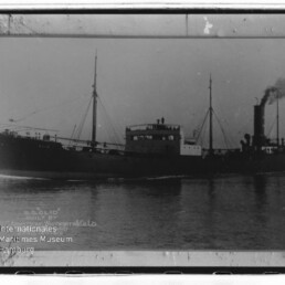 Schwarzweißfoto eines Dampfschiffs mit hohem Schornstein, zwei Masten und Rauch, auf ruhigem Wasser; Museumswasserzeichen links.