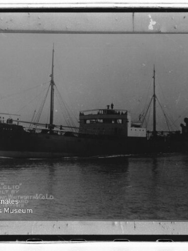 Schwarzweißfoto eines Dampfschiffs mit hohem Schornstein, zwei Masten und Rauch, auf ruhigem Wasser; Museumswasserzeichen links.