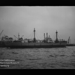 Schwarzweißbild eines Frachtschiffs mit Hafenkränen, Wasser im Vordergrund und Wasserzeichen Maritimes Museum Hamburg unten links.