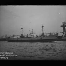 Schwarzweißfoto eines Frachtschiffs im Hafen mit Krananlage am rechten Bildrand und sichtbarem Museumslogo.