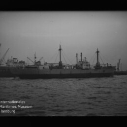 Frachtschiff mit Ladebäumen und Masten vor Hafenkränen, Wasser im Vordergrund und Museumswasserzeichen links unten.