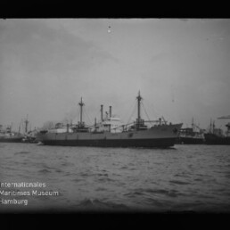 Frachtschiff im Hafen mit Kränen und anderen Schiffen im Hintergrund; sichtbares Museumslogo links unten.