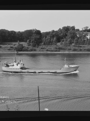 Kleiner Frachter auf ruhigem Fluss vor bewaldetem Ufer; Schwarzweißbild mit Maritimen-Museum-Wasserzeichen.