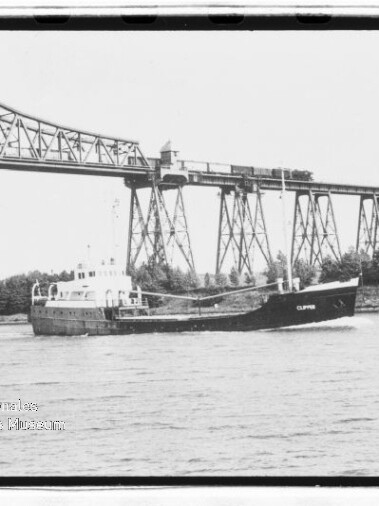 Frachtschiff 'Clipper' fährt auf dem Fluss vor einem hohen stählernen Eisenbahnviadukt mit Pylonen, Ufer mit Bäumen.