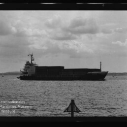 Frachtschiff mit Containern auf ruhiger See vor bewölktem Himmel; Museum-Logo Internationales Maritimes Museum Hamburg unten links.