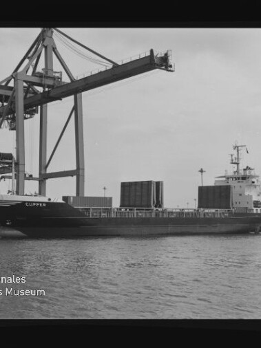 Frachtschiff CLIPPER am Kai unter einem großen Container-Kran, mehrere Container an Deck und Wasser im Vordergrund.