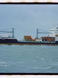 Frachtschiff mit Containern und zwei Ladekränen auf ruhiger See unter grauem Himmel; Museumslogo links unten.