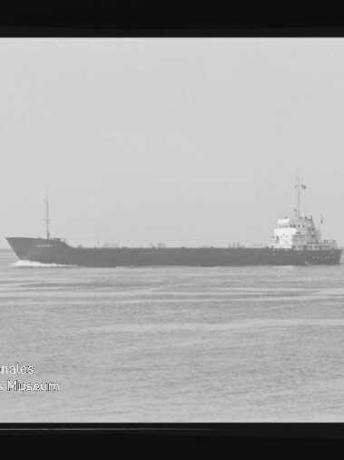 Frachtschiff in Seitenansicht auf ruhigem Meer vor flachem Horizont, schwarzweiß mit Museumswasserzeichen unten links.