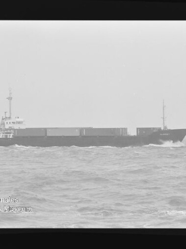 Schwarzweiß-Aufnahme eines Containerschiffs auf bewegter See, Filmrahmen und Museumslogo unten links sichtbar.