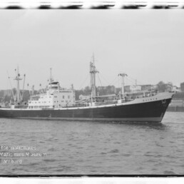 Schwarzweißaufnahme eines Frachtschiffs mit Ladebäumen im Hafen; links unten Stempel Internationales Maritimes Museum Hamburg.