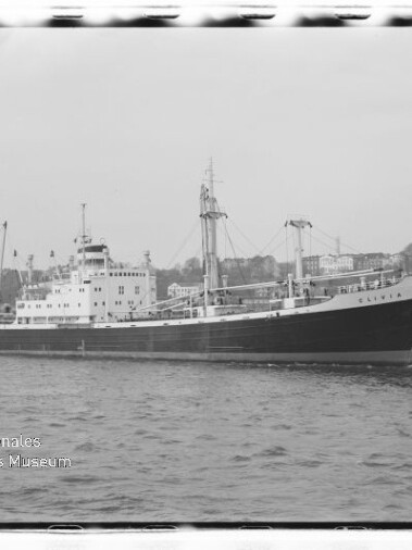 Schwarzweißaufnahme eines Frachtschiffs mit Ladebäumen im Hafen; links unten Stempel Internationales Maritimes Museum Hamburg.