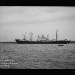 Großes Frachtschiff mit mehreren Ladebäumen auf ruhiger See, kleines Ruderboot im Vordergrund und Museumswasserzeichen unten links.