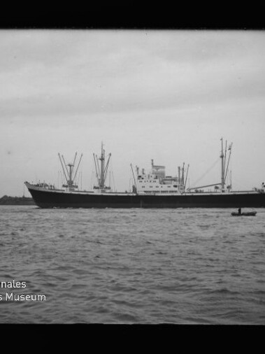 Großes Frachtschiff mit mehreren Ladebäumen auf ruhiger See, kleines Ruderboot im Vordergrund und Museumswasserzeichen unten links.