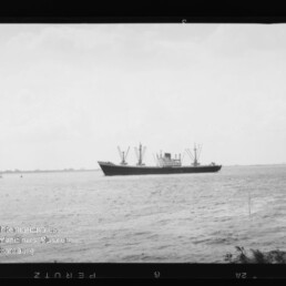 Frachtschiff mit mehreren Ladekränen auf ruhiger See, niedriger Horizont, kleines Uferstück links und leichter Wolkenhimmel