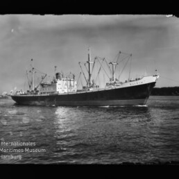 Graustufiges Frachtschiff mit Ladebäumen und Brückenaufbau auf ruhigem Wasser; Museumslogo unten links.