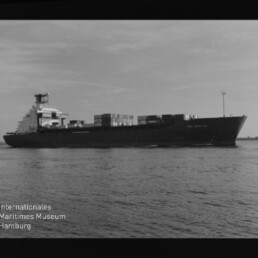 Containerschiff mit gestapelten Containern auf ruhigem Wasser, schwarzweiß, Museumslogo unten links