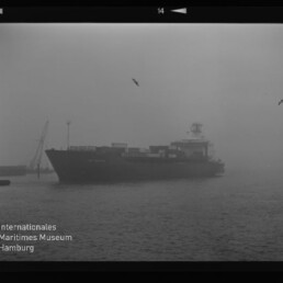 Containerschiff fährt durch dichten Nebel im Hafen, Container auf Deck, Möwen am Himmel und Museumslogo links unten