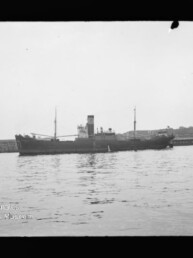 Frachtschiff mit mittigem Schornstein und zwei Masten vor Pier, spiegelnde ruhige Wasserfläche; Museumswasserzeichen links unten