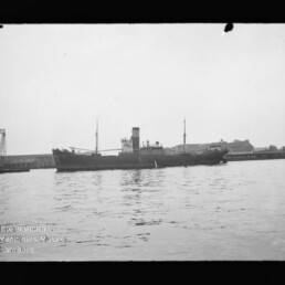 Frachtschiff mit mittigem Schornstein und zwei Masten vor Pier, spiegelnde ruhige Wasserfläche; Museumswasserzeichen links unten