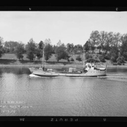 Kleiner Frachter fährt auf ruhigem Fluss vor baumbewachsenem Ufer, schwarzweißes Foto mit Filmrand und Museumswasserzeichen.