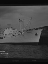 Weißes Schiff mit Namen CIMBRIA am Kai vor Lagerhäusern im Hafen unter bedecktem Himmel.