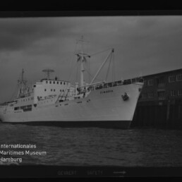 Weißes Schiff mit Namen CIMBRIA am Kai vor Lagerhäusern im Hafen unter bedecktem Himmel.