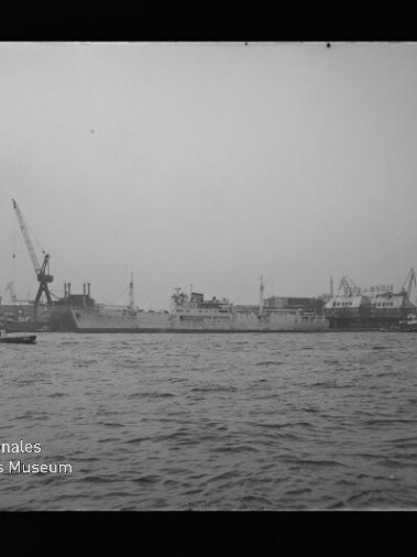 Großes Handelsschiff am Werftkai vor mehreren Hafenkränen, Wellengang im Vordergrund; kleines Boot links.