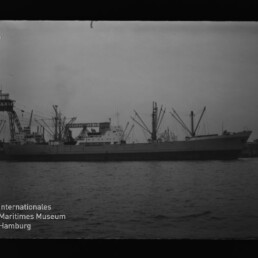 Großes Frachtschiff mit mehreren Ladekränen am Kai, ruhiges Hafenwasser und Museumsschriftzug unten links.