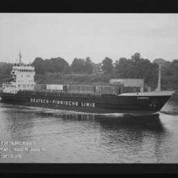 Frachtschiff Cimbria der Deutsch‑Finnischen Linie fährt auf ruhigem Wasser, Container an Deck, Baumufer im Hintergrund.