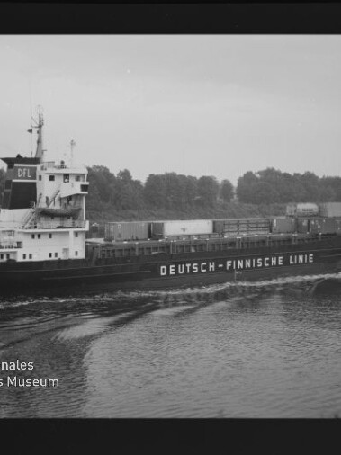 Frachtschiff mit Aufschrift DEUTSCH-FINNISCHE LINIE auf ruhigem Wasser vor einer Baumreihe; Schwarzweißaufnahme mit Museumslogo.