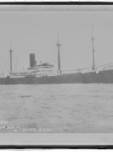 Schwarzweißfoto eines Frachtdampfers mit Mittelschornstein und drei Masten auf ruhigem Wasser; Museumswasserzeichen links unten.