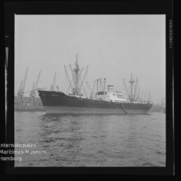 Schwarzweißfoto eines Frachtschiffs an einem Hafen mit Kränen im Hintergrund und Museumslogo unten links.