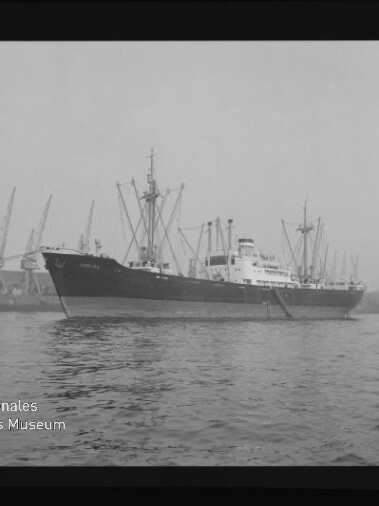 Schwarzweißfoto eines Frachtschiffs an einem Hafen mit Kränen im Hintergrund und Museumslogo unten links.