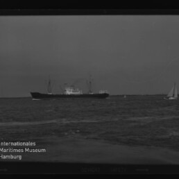 Frachtschiff mittig auf ruhiger See, kleines Segelboot rechts und Wasserzeichen Internationales Maritimes Museum links.