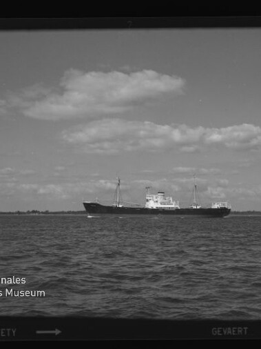 Frachtschiff auf ruhiger See vor flachem Küstenhorizont unter wolkigem Himmel; Logo des Maritimen Museums Hamburg sichtbar.