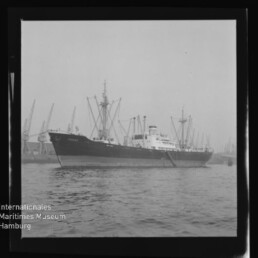 Frachtschiff mit Ladegeschirr auf dem Wasser vor Hafenkränen; Museumslogo und Schriftzug unten links.