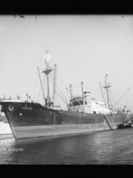 Frachtschiff Coburg am Kai eines Hafens mit Ladekränen an Bord und einem Schlepper längsseits.