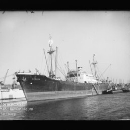 Frachtschiff Coburg am Kai eines Hafens mit Ladekränen an Bord und einem Schlepper längsseits.