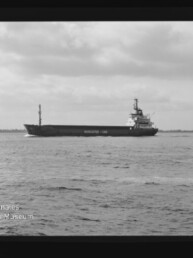 Schwarzweißaufnahme eines Frachtschiffs auf ruhiger See unter bewölktem Himmel, eingerahmt von sichtbarem Filmrand.