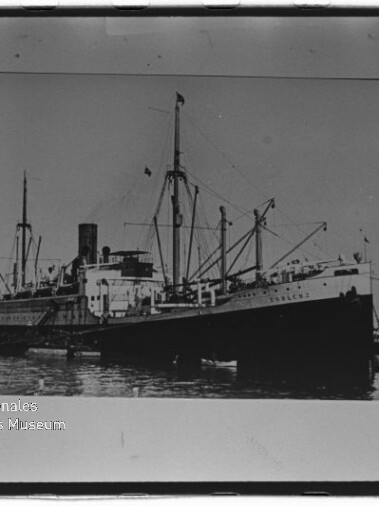 Schwarzweiß-Frachtdampfer mit Schornstein, Masten und Ladebäumen im Hafen; Museumswasserzeichen unten links.