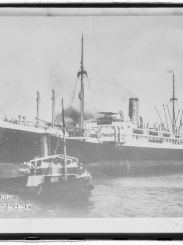 Großes Dampfschiff am Kai mit vorausfahrendem Schlepper, Rauch aus dem Schornstein und Hafenbauten im Hintergrund.