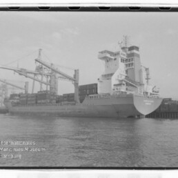 Containerschiff am Kai mit Containern an Deck und großen Hafenkränen im Hintergrund; Museumslogo links unten.